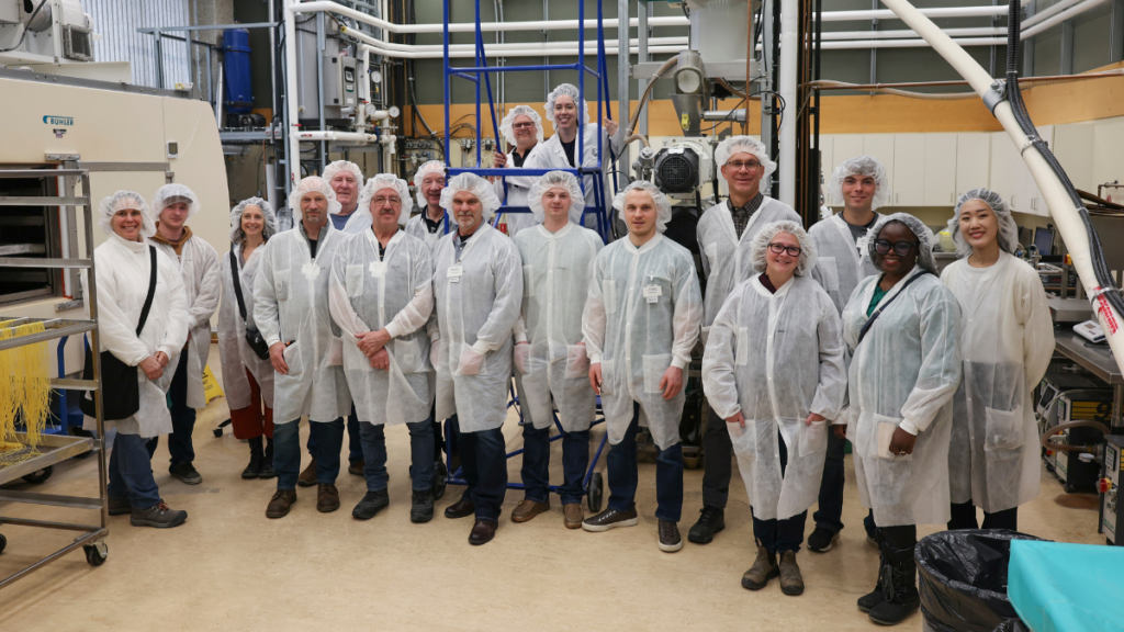 A group of farmers wearing lab coats and hairnets pose for a photo in a pasta manufacturing and testing facility.